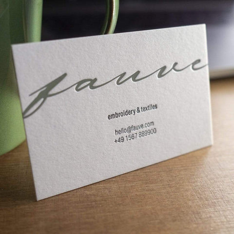 Eine geprägte Letterpress Visitenkarte mit feiner Haptik und spürbarem Druckbild. Zweifarbiger Druck in grün-grau, das Logo, und schwarz, der Text, auf naturweißem Papier. Beispiellogo in Handschrift "fauve" mit Untertitel "embroidery & textiles" und E-Mail-Adresse und Telefonnummer.