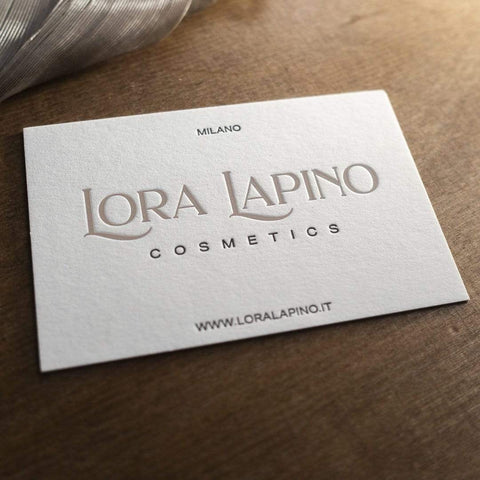 Eine geprägte Letterpress Visitenkarte mit feiner Haptik und spürbarem Druckbild. Zweifarbiger Druck in beige-gold, das Logo, und schwarz, der Text, auf naturweißem Papier. Beispiellogo "LORA LAPINO COSMETICS" mit Ortsangabe und Internetseite.